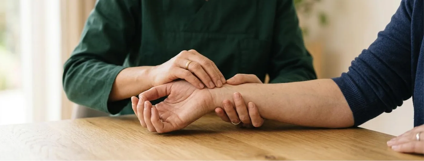 Karpaltunnelsyndrom: Zwei Therapeutinnenhände untersuchen behutsam das Handgelenk einer erwachsenen Person auf einem Holztisch (Symbolbild, mit künstlicher Intelligenz erzeugt)